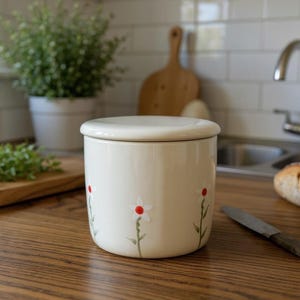 May include: A cream ceramic butter dish with a lid, decorated with red and white floral accents. The dish is on a wooden countertop, with a loaf of bread and a knife nearby. A cutting board with herbs is also visible.
