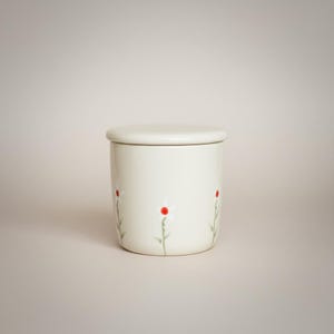 May include: A cream-coloured ceramic jar with a matching lid. The jar is decorated with a simple daisy pattern, featuring white petals and red centres on green stems. The jar is cylindrical and sits on a neutral background.