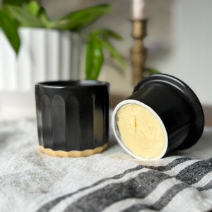 May include: Two black ceramic butter dishes with a gold rim. One dish is filled with a block of yellow butter.