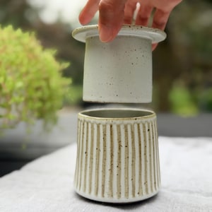Op de afbeelding: Een keramische boterpot bestaande uit twee delen: een gespikkeld gebroken wit klokvormig deksel en een bijpassende basis met een verticale ribbelstructuur. Het deksel wordt boven de basis gehouden, wat de gespikkelde afwerking toont.