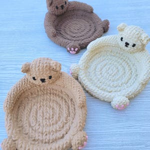May include: Three crocheted bear-shaped trinket dishes in brown, beige, and white. Each dish has a small pink paw print on each side.