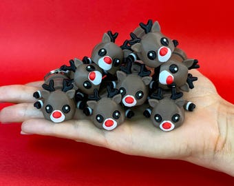May include: A collection of brown reindeer figurines with black antlers, white eyes, and red noses. The reindeer are small and have a cartoonish design. The figurines are held in a hand against a red background. Christmas-themed decorations.