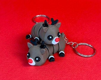May include: A keychain featuring two brown reindeer figurines with red noses. The reindeer are stacked on top of each other, with the top reindeer's head tilted back. The keychain is made of metal and has a silver finish.
