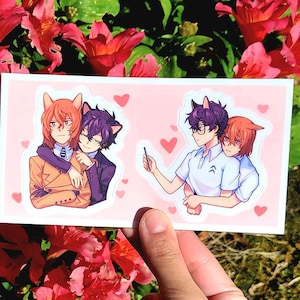 May include: A rectangular sticker featuring two pairs of anime-style characters with cat ears, embracing each other. The characters are depicted in different outfits, with hearts scattered around them. The sticker is held against a backdrop of red flowers and green foliage.