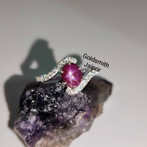 Peut inclure: Une bague en argent avec une pierre précieuse en rubis étoilé rouge et de petits diamants clairs sertis dans une bande incurvée. La bague est posée sur une roche violette. Goldsmith Jaipur.