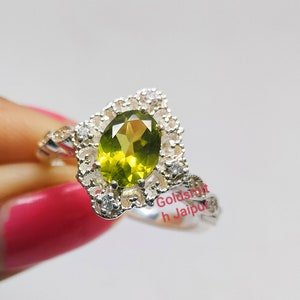 Könnte beinhalten: Ein silberner Ring mit einem grünen ovalen Edelstein, der von kleineren weißen Edelsteinen umgeben ist. Der Ring hat ein zartes, filigranes Design.