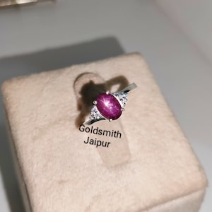Puede incluir: Un anillo de plata con una gema grande, ovalada, de color rojo intenso con efecto estrella. El anillo tiene un diseño de caña dividida y está adornado con pequeñas piedras transparentes. El texto "Goldsmith Jaipur" es visible.