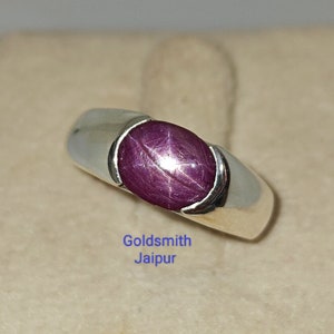Puede incluir: Un anillo de plata con una piedra preciosa de rubí estrellado rojo oscuro. El anillo está engastado en una banda simple con un acabado liso. El texto "Goldsmith Jaipur" es visible debajo del anillo.
