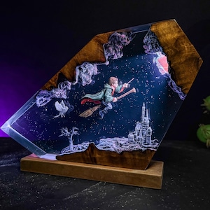 May include: A decorative resin art piece featuring a wizard on a broomstick flying through a starry night sky. The artwork includes a castle, a bird, and a tree, all set within a wooden frame and base. The piece is illuminated with a soft, internal light.