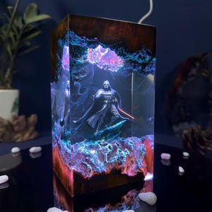 May include: A dark blue and purple resin lamp with a Darth Vader figurine inside. The lamp is lit from the inside, creating a glowing effect. The resin base is textured and has a dark brown color.