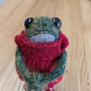 May include: A knitted frog ornament with large black eyes and a red scarf. The frog's body is green with yellow flecks, and the scarf is a deep red. A charming, handcrafted item.