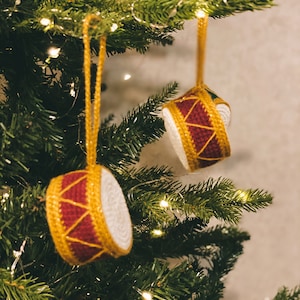 Puede incluir: Dos adornos navideños de crochet con forma de tambor. Los tambores son rojos y dorados con detalles blancos. Están colgados de un árbol de Navidad.
