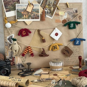 May include: A craft display featuring a collection of miniature crocheted sweaters, scarves, and a sorting hat. Vintage postcards and a camera are arranged on a desk with various craft supplies, including twine, scissors, and rolled paper.