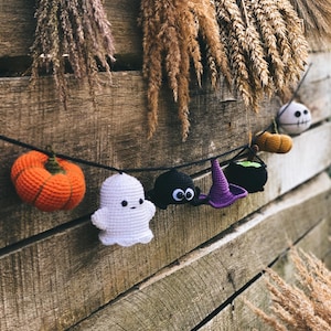 Op de afbeelding: Een Halloween-krans met gehaakte decoraties, waaronder een pompoen, een spook, een spin en een heksenhoed. De krans hangt aan een houten achtergrond met gedroogde grassen.