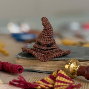May include: A brown crocheted Sorting Hat, a miniature replica, sits atop a vintage book. The hat's pointed crown and wide brim are clearly defined. A red and gold striped scarf and a red crocheted item are in the foreground.