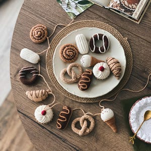Könnte beinhalten: Eine Girlande aus gehäkelten Backwaren und Desserts, darunter Donuts, Brezeln und Eiswaffeln, die auf einem weißen Teller angeordnet sind. Die handgefertigten Artikel sind in Braun-, Beige- und Weißtönen gehalten, mit roten Akzenten.