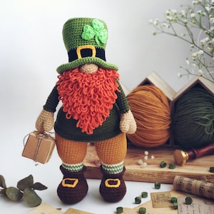 Puede incluir: Un gnomo de crochet que representa a un duende, con un sombrero verde con un trébol, una camisa verde y naranja, pantalones naranjas y zapatos marrones. El gnomo lleva una pequeña caja marrón.