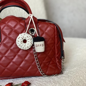 Pode incluir: Uma bolsa de mão acolchoada vermelha com uma alça de corrente prateada e um amuleto de donut e xícara de café de crochê branco.