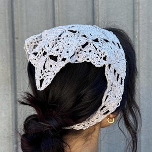 May include: A white crocheted headband with a delicate floral pattern. The headband is worn on a person with dark hair pulled back in a bun.