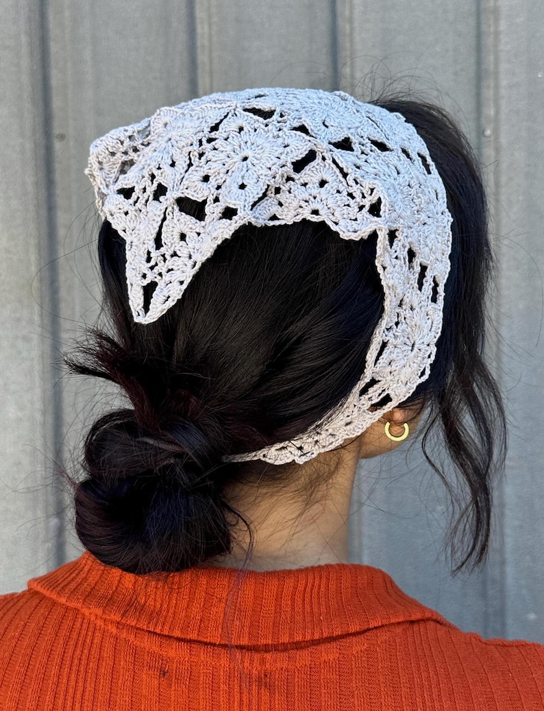 May include: A white crocheted headband with a delicate floral pattern. The headband is worn on a person with dark hair pulled back in a bun.