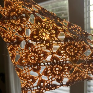 May include: A close-up of a crocheted lace doily with a sunbeam shining through it. The doily is a light brown color and has a delicate, intricate pattern.