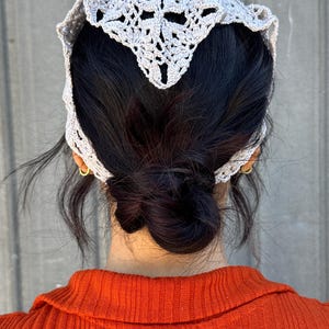 May include: A gray crocheted hair scarf with a delicate pattern. The scarf is worn on a person with dark hair pulled back in a bun.
