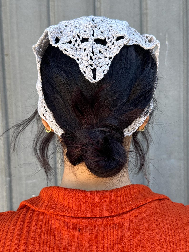 May include: A gray crocheted hair scarf with a delicate pattern. The scarf is worn on a person with dark hair pulled back in a bun.