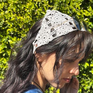 May include: A woman with long, dark hair is wearing a light gray crocheted headband. The headband is tied in a knot at the back of her head. She is wearing a blue shirt and has a pair of round sunglasses on top of her head.