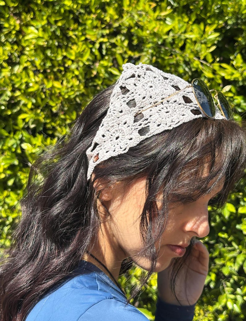 May include: A woman with long, dark hair is wearing a light gray crocheted headband. The headband is tied in a knot at the back of her head. She is wearing a blue shirt and has a pair of round sunglasses on top of her head.