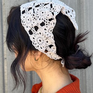 May include: A light gray crocheted bandana with a floral pattern. The bandana is tied around the head of a person with dark hair.
