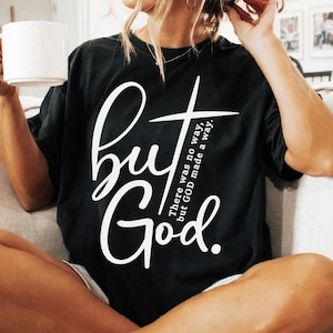 May include: Black t-shirt with the words "but God" in a white script font, with a cross graphic. The shirt also has the text "There was no way, but GOD made a way." A white mug is held in the person's hand.