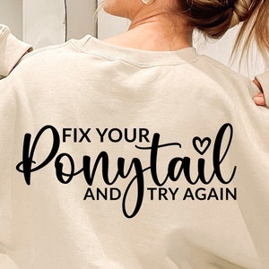 Può includere: Una felpa beige chiaro con testo nero che recita "Fix your ponytail and try again" con un piccolo simbolo di cuore.