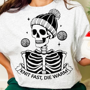 May include: A gray t-shirt with a black and white illustration of a skeleton wearing a knit hat and holding yarn balls. The text "Knit Fast, Die Warm" is printed on the shirt.