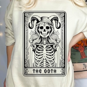 以下が含まれることがあります： 黒い枠の中に、角のある骸骨の白黒イラストと「The Goth」という文字が書かれた、ベージュのトレーナーです。