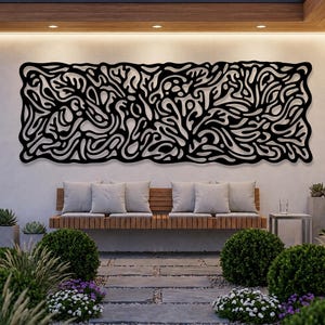 May include: A large, black metal wall art piece with an abstract, organic design is mounted on a white wall. The artwork is above a wooden bench with gray cushions. The outdoor setting includes greenery, stone pavers, and a modern architectural design.