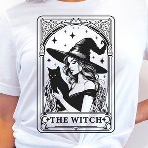 Puede incluir: Camiseta blanca con una ilustración en blanco y negro de una bruja sosteniendo un gato negro. La ilustración está enmarcada en un borde decorativo y el texto "THE WITCH" está debajo de la imagen.