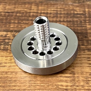 May include: A silver metal spinning top with a round base and a central spindle with a textured surface. The base has a ring of small holes around the spindle.