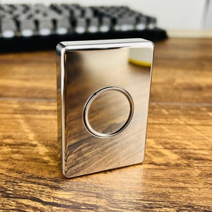 May include: A silver metal fidget toy with a round indentation in the center. The toy is on a wooden surface.