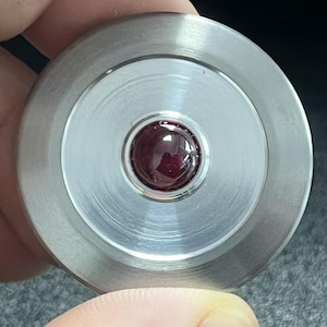 Puede incluir: Un disco circular de metal plateado cepillado con un acabado liso y pulido. En el centro del disco hay una pequeña piedra preciosa redonda de color rojo intenso, engastada de forma segura en la superficie metálica, creando un contraste marcado.