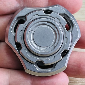 May include: A silver metal fidget spinner with a gear design. It measures 48x48x17 mm and weighs 112 grams.