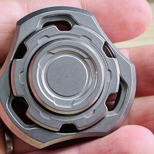 May include: A silver metal fidget spinner with a circular design and multiple rings. The spinner has a textured surface and a geometric shape.