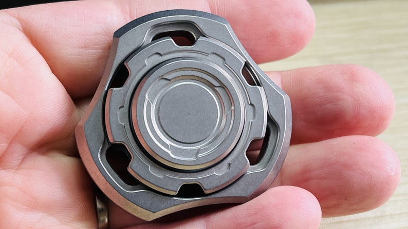 May include: A silver metal fidget spinner with a circular design and multiple rings. The spinner has a textured surface and a geometric shape.