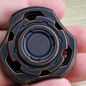 May include: A copper-colored fidget spinner with a complex, multi-layered design. The spinner has a central circle with a ring around it, and the outer edges are shaped like a star.