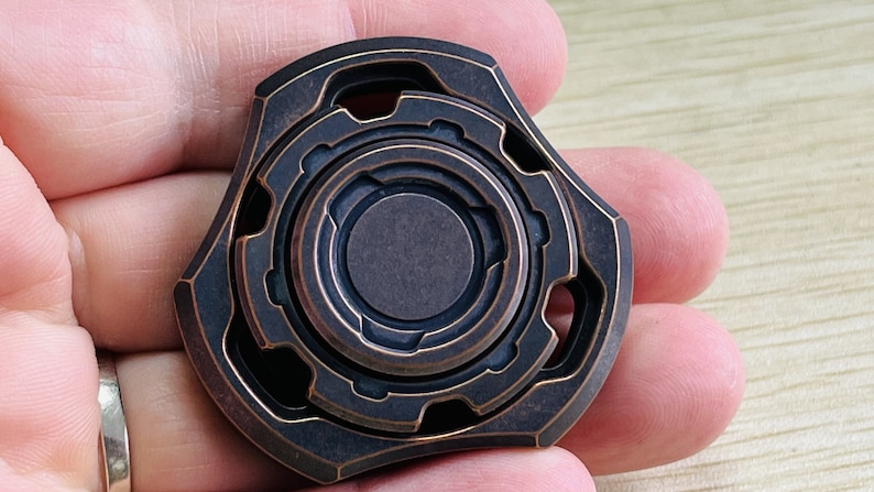 May include: A copper-colored fidget spinner with a complex, multi-layered design. The spinner has a central circle with a ring around it, and the outer edges are shaped like a star.