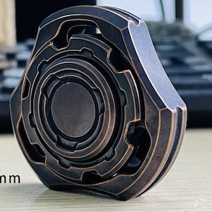 May include: A bronze fidget spinner with intricate geometric designs. It measures 48x48x17 mm and weighs 127 grams.