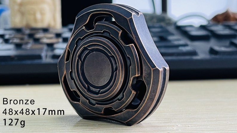 May include: A bronze fidget spinner with intricate geometric designs. It measures 48x48x17 mm and weighs 127 grams.