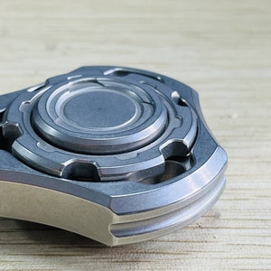 May include: A silver metal fidget spinner with a multi-layered design. The spinner has a central disc with a raised outer edge and a series of smaller, concentric rings.