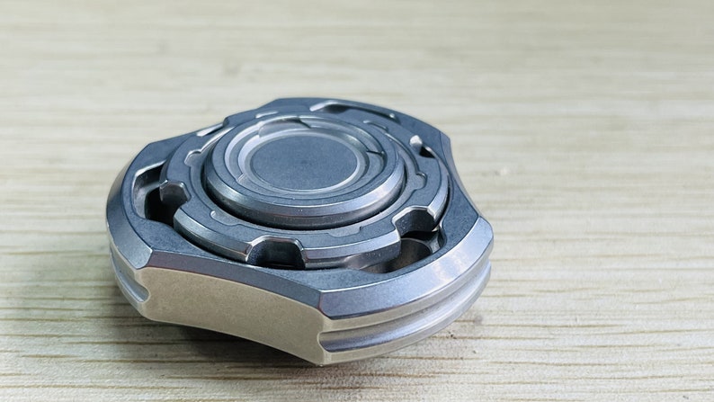 May include: A silver metal fidget spinner with a multi-layered design. The spinner has a central disc with a raised outer edge and a series of smaller, concentric rings.