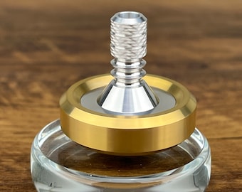 May include: A gold and silver spinning top on a clear glass base. The top has a textured silver cap and a gold ring around the base.