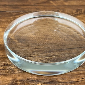 May include: A clear, circular glass object with a flat base and rounded edges. The transparent disc is approximately 15 cm in diameter and appears to be made of thick glass. The object is likely a decorative piece or a functional item.